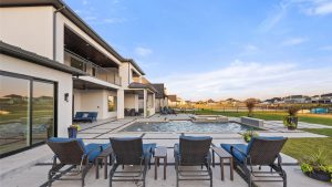Modern backyard with a swimming pool, lounge chairs, and a spacious patio area next to a two-story house. Green lawn and neighboring houses are visible in the background under a blue sky.