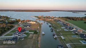 Aerial view of a lakeside neighborhood with houses along canals leading to a large lake. A red location marker highlights one house near the water’s edge. Lush green lawns and docks are visible.