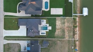 Aerial view of a lakeside property with a large house, central backyard pool, paved patio, driveway, and a covered dock extending over the water. The lot is outlined in white. Neighboring homes and lawns are visible.