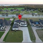 Aerial view of a suburban neighborhood with houses along a canal. A red location pin is placed above the roof of one house, indicating it as a specific property. Trees and grassy areas are visible in the background.
