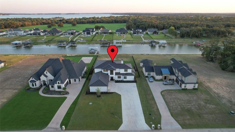 Aerial view of a suburban neighborhood with houses along a canal. A red location pin is placed above the roof of one house, indicating it as a specific property. Trees and grassy areas are visible in the background.