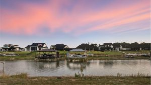 A serene lakeside scene at sunset with modern houses, private docks, and boats along the shore; pastel pink and purple clouds fill the sky, reflecting peacefully on the water.