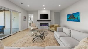 Modern living room with light walls and a large sectional sofa, glass coffee table, fireplace under a wall-mounted TV, beige armchair, and a colorful fish painting. Sliding glass doors open to a patio area.