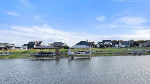 Waterfront homes with boat docks and covered patios line a grassy shoreline under a blue sky with scattered clouds. The calm water reflects the houses and docks.