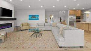 A modern living room with a large gray sectional sofa, glass coffee table, beige armchair, wall-mounted TV, electric fireplace, and open kitchen with light wood cabinets and stainless steel appliances. A fish artwork hangs on the wall.