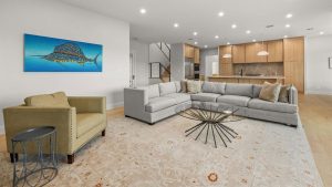 Modern living room with a large light gray sectional sofa, a green armchair, a glass coffee table, and a patterned rug. An abstract fish painting hangs on the wall, and an open kitchen is visible in the background.