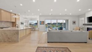 Bright open-plan living area with a modern kitchen, large island, light wood cabinets, dining table, gray sectional sofa, fireplace, and floor-to-ceiling windows overlooking a patio and waterfront view.