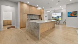 Modern kitchen with light wood cabinets, a large marble island with a built-in sink, pendant lights, light wood floors, and an adjacent dining area with large windows showing a view outside.