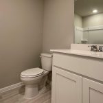 A modern bathroom with light-colored walls, a white toilet, a white vanity with a sink and faucet, a large mirror, and a shower in the background. The floor has light wood-like tiles.