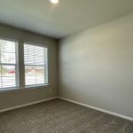 Small, empty room with beige carpet, light gray walls, and a large window with white blinds letting in daylight. Ceiling light is on, and room appears clean and unfurnished.