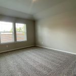 Empty room with beige walls, a light gray carpet, and two large windows letting in natural light. The windows show a view of a brick wall and some greenery outside. The room appears clean and spacious.