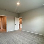 Empty room with beige carpet, light gray walls, a ceiling light, and two white doors—one open to a hallway with wood flooring, and the other to another area of the house. Natural light fills the space.