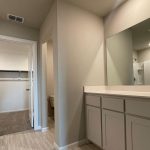 Modern bathroom with a large mirror, double sink vanity, light wood flooring, and beige walls. An open door reveals a walk-in closet with shelves and carpeted floor. Another doorway leads to a toilet area.