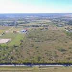 Aerial view of open grassy land with scattered bushes, a few buildings, a road lined with trees in the foreground, and suburban neighborhoods visible in the distance under a clear blue sky.