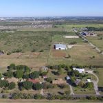 Aerial view of a rural area with scattered houses, trees, open fields, and a large barn. Roads run through the landscape, and a city is visible on the distant horizon under a clear blue sky.
