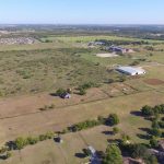 Aerial view of a rural landscape with open fields, scattered trees, a few houses, and farm buildings under a clear sky. Residential areas and distant developments are visible in the background.