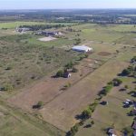 Aerial view of a rural landscape featuring open green fields, scattered trees, a few buildings, and a residential area with houses lining a road on the right side. The horizon stretches under a clear blue sky.