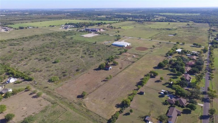 Aerial view of a rural landscape featuring open green fields, scattered trees, a few buildings, and a residential area with houses lining a road on the right side. The horizon stretches under a clear blue sky.