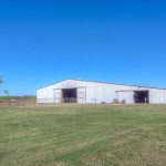 A large white metal barn stands on a flat grassy field under a clear blue sky, with a few small trees and fences nearby. The area appears open and rural.