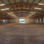 A large, empty indoor arena with a dirt floor, metal roof, and metal walls. Sunlight shines in through the open end, revealing a view of green trees outside.
