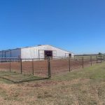 A large white barn with a metal roof sits in an open grassy field, surrounded by fenced dirt paddocks under a clear blue sky. A horse stands in one of the fenced areas to the left of the barn.