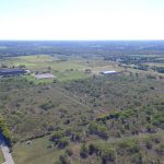 Aerial view of a rural landscape with open fields, scattered trees, and a few buildings along a road. The horizon stretches out under a clear sky, showing distant farmland and greenery.
