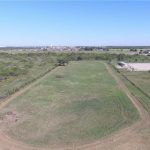 Aerial view of a large, open grassy field with a dirt road curving around it, sparse trees surrounding the area, and buildings visible in the distance under a clear blue sky.