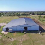 A large metal barn or warehouse with a gray roof sits in the middle of a grassy, rural landscape with scattered trees and dirt paths under a clear sky. Farmland and fields stretch into the distance.