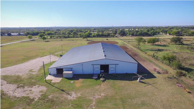 A large metal barn or warehouse with a gray roof sits in the middle of a grassy, rural landscape with scattered trees and dirt paths under a clear sky. Farmland and fields stretch into the distance.
