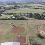 Aerial view of a rural landscape with fields, scattered trees, dirt arenas, fenced areas, a few buildings, and a road running through the scene under a clear sky.