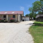 A stone house with a red metal roof sits beside a gravel driveway. There are rose bushes in front, green grass and trees to the side, and a metal barn structure in the background under a clear blue sky.