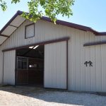 A light-colored metal barn with a brown roof and trim stands under a clear blue sky. The barn door is open, revealing horse stalls inside. A gravel area is in front, and tree branches partially frame the top left corner.