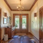 A rustic hallway with wooden doors, a cowhide rug, wooden furniture, antler chandelier, framed art on the wall, and double glass doors letting in natural light from outside.
