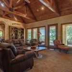 A spacious living room with leather sofas, wooden beams on a vaulted ceiling, large windows overlooking greenery, built-in bookshelves, and rustic decor. Natural light fills the room, highlighting wood and stone accents.