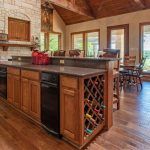 Spacious kitchen with wooden floors, a large island featuring a built-in wine rack and dishwasher, stone range hood, wood cabinets, and large windows letting in natural light with views of greenery outside.