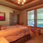 A cozy bedroom with a brown bed, wooden furniture, a TV, and large windows. The decor includes a Texas flag on the wall and star-shaped ornaments, creating a rustic, Southwestern feel. Sunlight streams through the window.