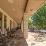 A spacious stone patio with rocking chairs, tables, and chairs under a covered roof, overlooking a lush green yard and swimming pool, shaded by large trees on a sunny day.