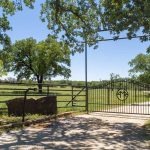 A metal ranch gate with a circular emblem stands open at the entrance to a rural property, surrounded by green grass, large trees, and a gravel driveway under a clear blue sky.