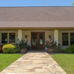 A stone walkway leads to the entrance of a house with a tan brick exterior, wooden double doors, and large windows. Neatly trimmed bushes, flowering plants, and trees line the path, with two decorative horse-head posts at the front.