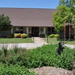 A single-story house with a brown roof and stone exterior sits behind a curved driveway and a well-maintained garden with green shrubs, flowering plants, and a black metal deer sculpture. Trees frame the property under a clear blue sky.