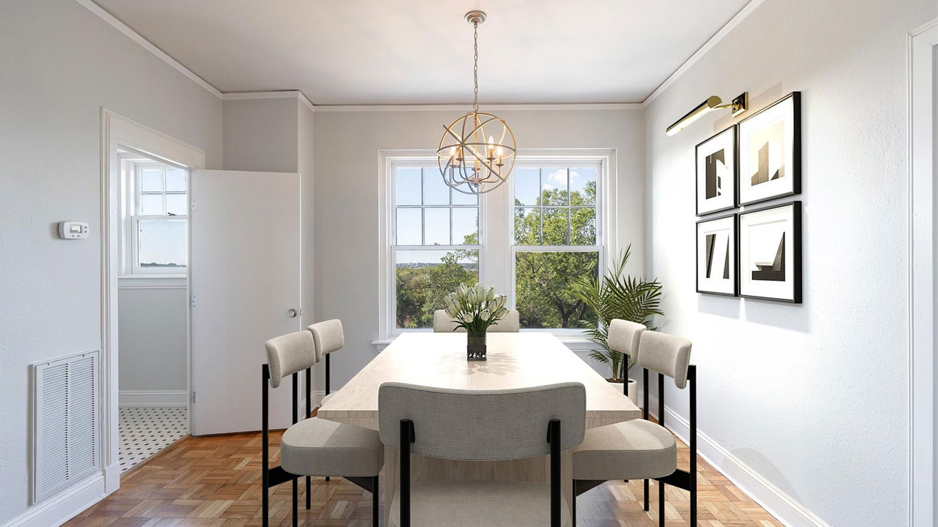 Bright dining room with a rectangular table, six upholstered chairs, a modern pendant light, framed abstract art on the wall, and two large windows with a view of green trees.