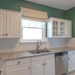 A kitchen with white cabinets, granite countertops, a stainless steel dishwasher, white subway tile backsplash, green walls, and a window above the sink with a beige shade.