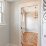 A bright hallway with black-and-white patterned tile flooring leads to a room with parquet wood floors, white walls, large windows, and a round, modern chandelier. Natural light fills both spaces.
