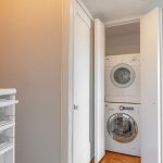 A compact laundry closet with white folding doors contains a stacked washer and dryer. The floor is wood parquet, and there are white storage shelves visible on the left side. The walls and ceiling are painted white.