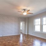 An empty room with light-colored walls, large windows, a ceiling fan with a light, and parquet wood flooring. Natural light fills the space, and a patterned accent wall is visible on one side.