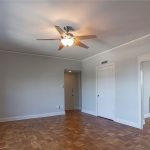 A bright, empty room with light gray walls, parquet wood floors, large windows, a ceiling fan with light, and white doors leading to adjacent spaces. Natural light fills the space.