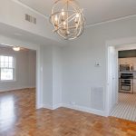 A bright, empty room with light gray walls, wooden parquet flooring, a modern chandelier, and a view into a kitchen with white cabinets and appliances. Large windows let in natural light.