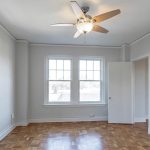 Bright, empty room with light gray walls, parquet wood floors, large windows letting in natural light, a ceiling fan with lights, and an open white door.