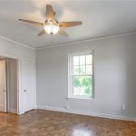 Empty room with light gray walls, wood parquet flooring, a ceiling fan with a light, two white doors, a window with green trees visible outside, and natural light coming through another large window.