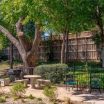 A fenced patio area with a round stone table and benches, metal chairs, and a grill is shaded by large trees. A wooden privacy fence and greenery are in the background.